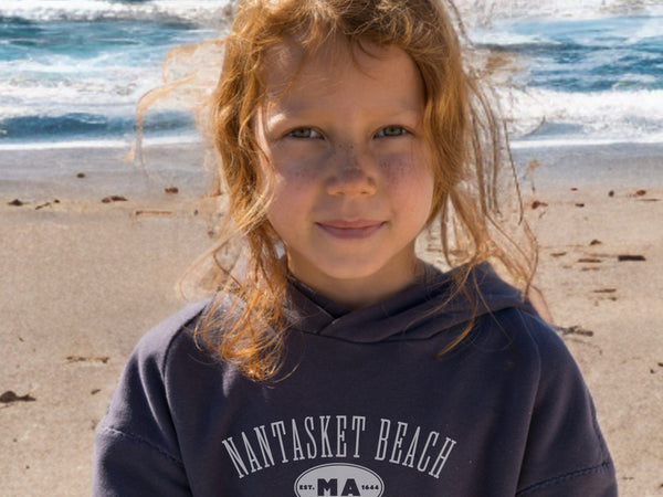 Young girl with sandy face and hair standing on a beach wearing a hoodie with 'Nantasket Beach, MA' text.