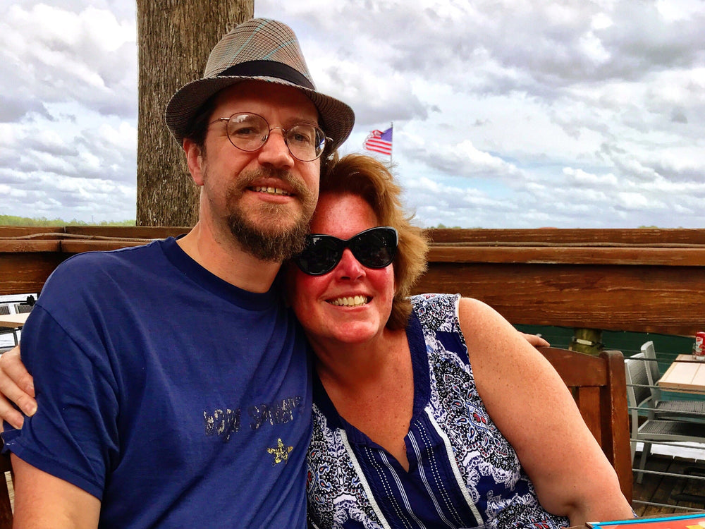 Two people sitting together outdoors with a cloudy sky and wooden structure in the background.