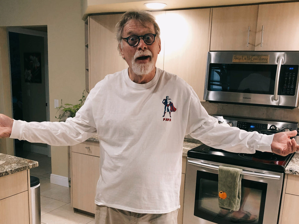 Man in a kitchen wearing a white long-sleeve shirt with a logo, standing with arms outstretched.