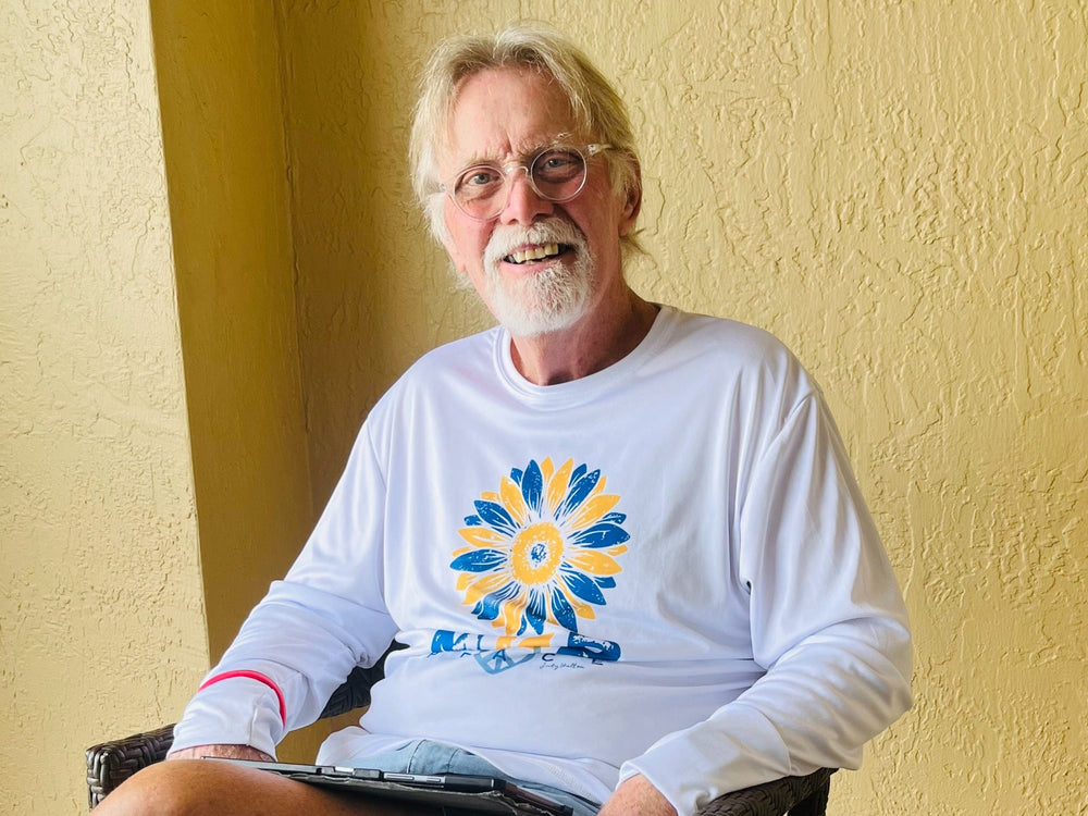 Man wearing a white long-sleeve shirt with a colorful design, sitting against a yellow wall.