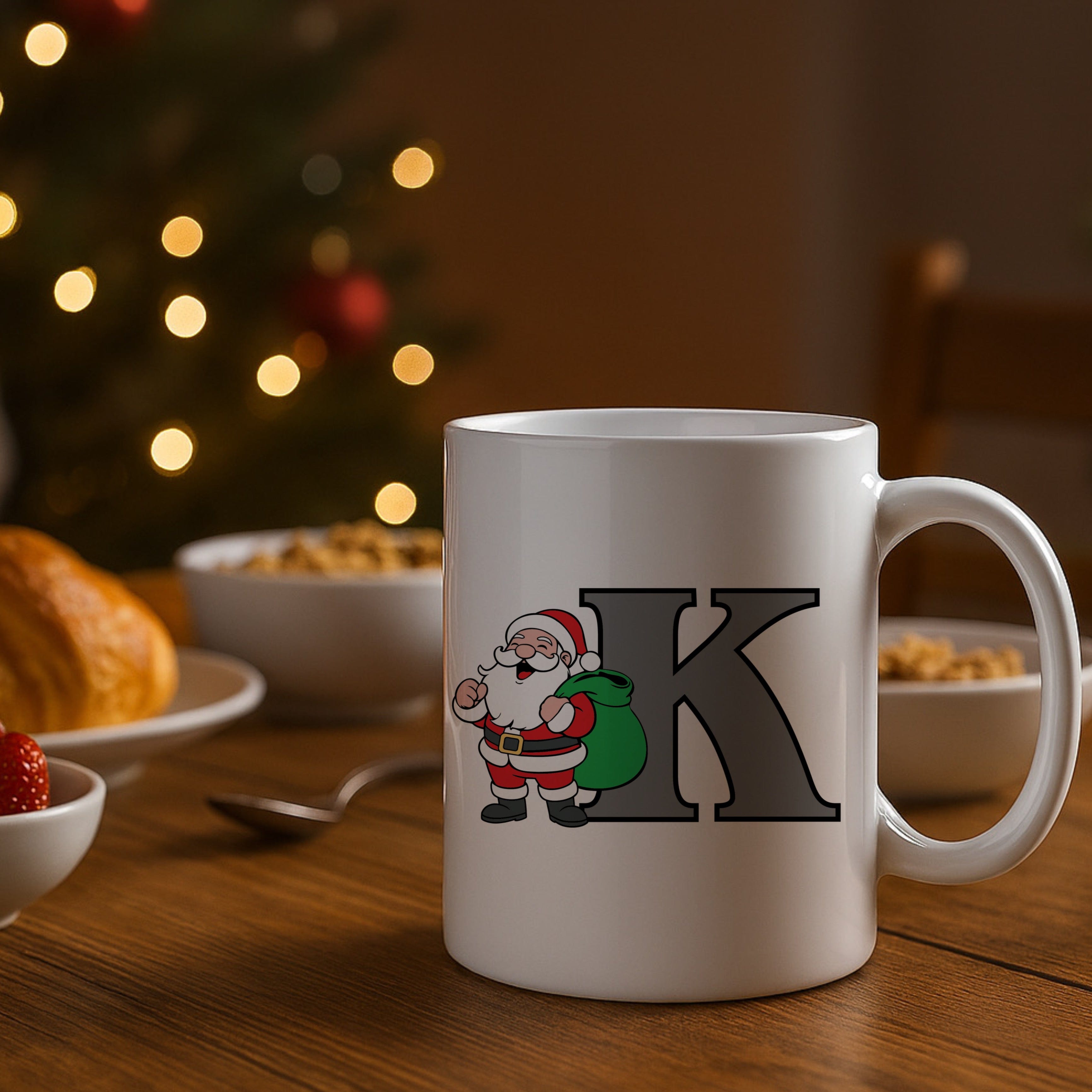 White mug with Santa Claus design and letter 'K' on a table with a Christmas tree in the background.