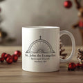White mug with 'St. John the Evangelist Episcopal Church' design on a wooden surface with Christmas decorations.